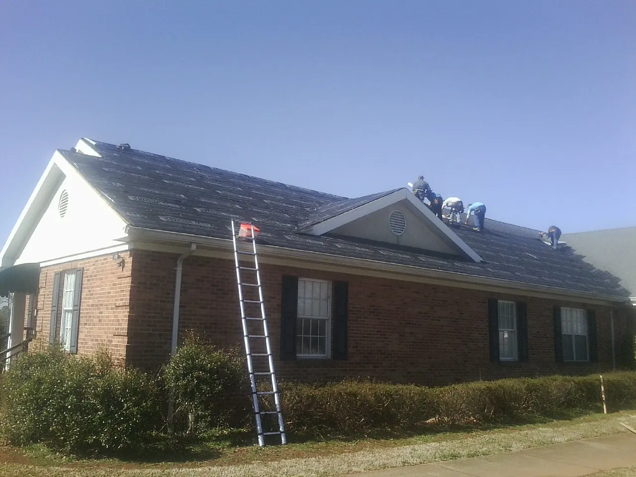 Crew tearing shingles off a brick ranch roof with a ladder set — Metal Roof Repair work by our crew in Village Mills