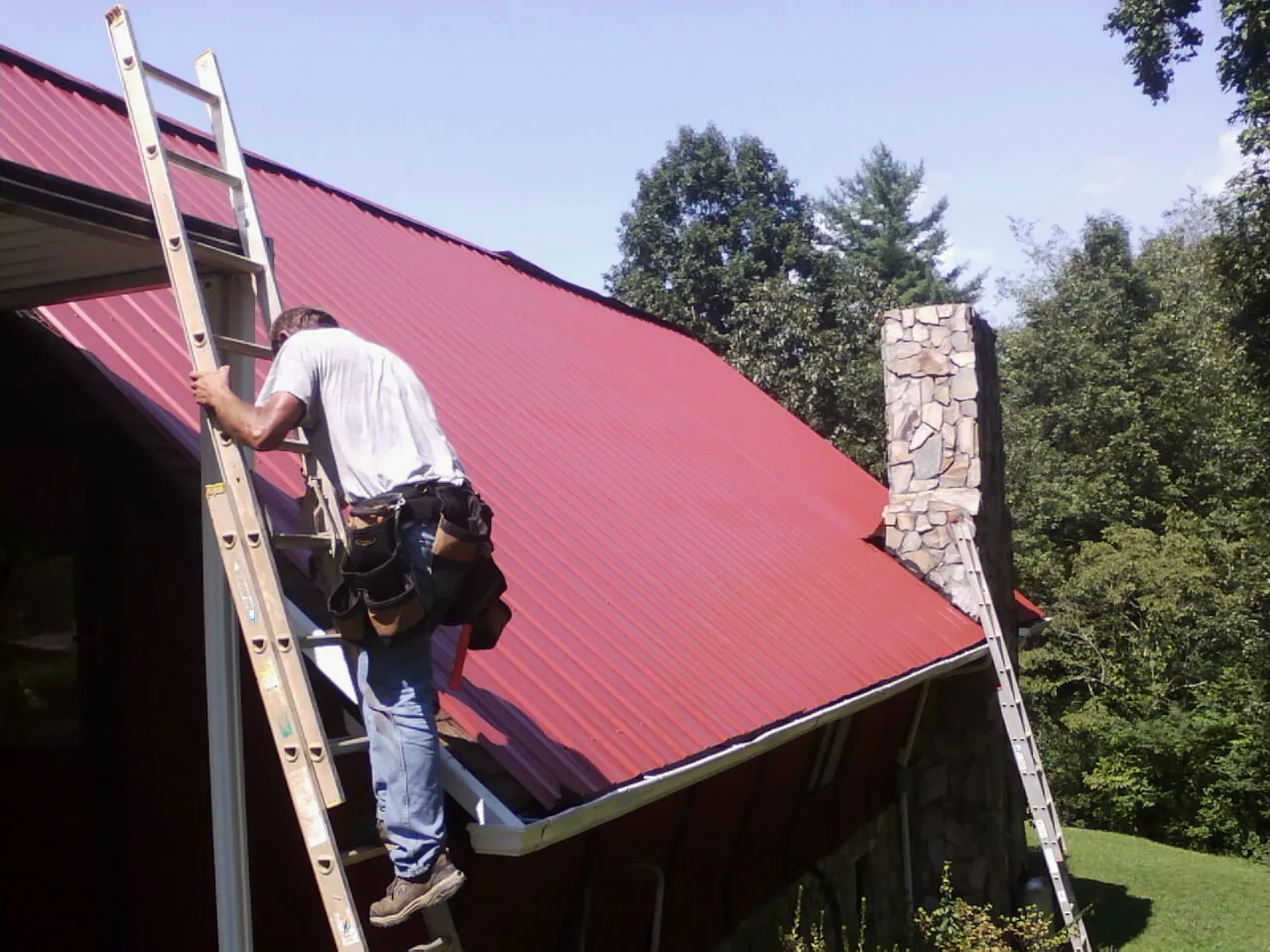 Roofer on a ladder beside a red metal roof and stone chimney — Soffit & Fascia Repair by Roofing Brothers in Village Mills